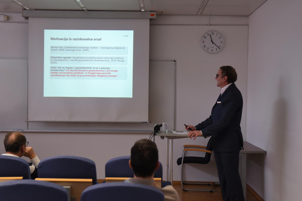 Oseba v obleki predstavi diaprojekcijo v učilnici, udeleženci pa sedijo. Besedilo na diapozitivih je v tujem jeziku. Na uri je 9:10.