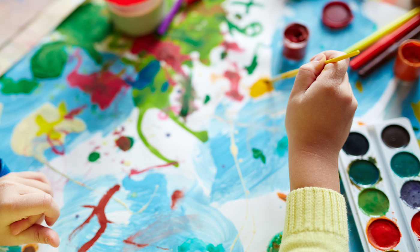 Otroška roka ustvarja barvito sliko z vodenimi barvami. Slika predstavlja ustvarjanje za razstavo otroških likovnih del »Denarček«.