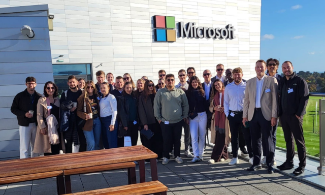 Raznolika skupina ljudi se smeji pod jasnim modrim nebom. Stojijo pred logotipom Microsoft na sodobni stavbi.