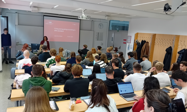 Učilnica, polna študentov, ki sedijo v vrstah in so osredotočeni na predavanje. Na velikem zaslonu spredaj je prikazana predstavitev o navajanju virov po APA.