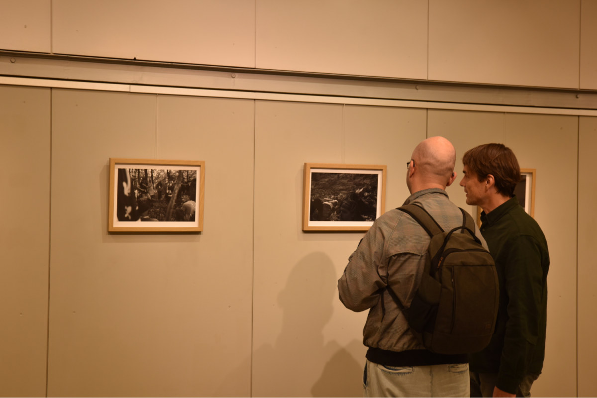 Dva moška stojita blizu galerijske stene in natančno pregledujeta črno-bele fotografije v lesenih okvirjih. Eden od njiju nosi nahrbtnik, kar ustvarja zanimivo vzdušje.