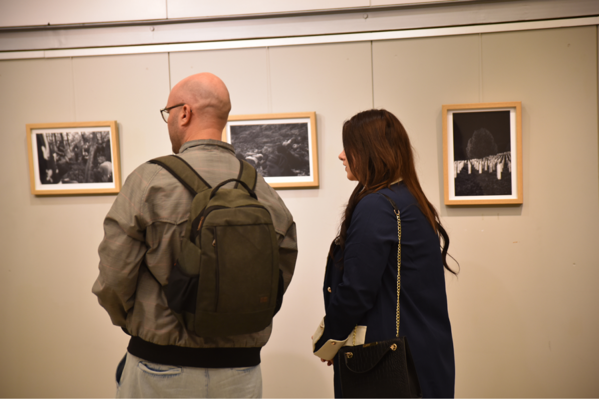 Dve osebi, moški z nahrbtnikom in ženska z torbico, stojita v galeriji in si ogledujeta tri črno-bele fotografije, ki so razstavljene.
