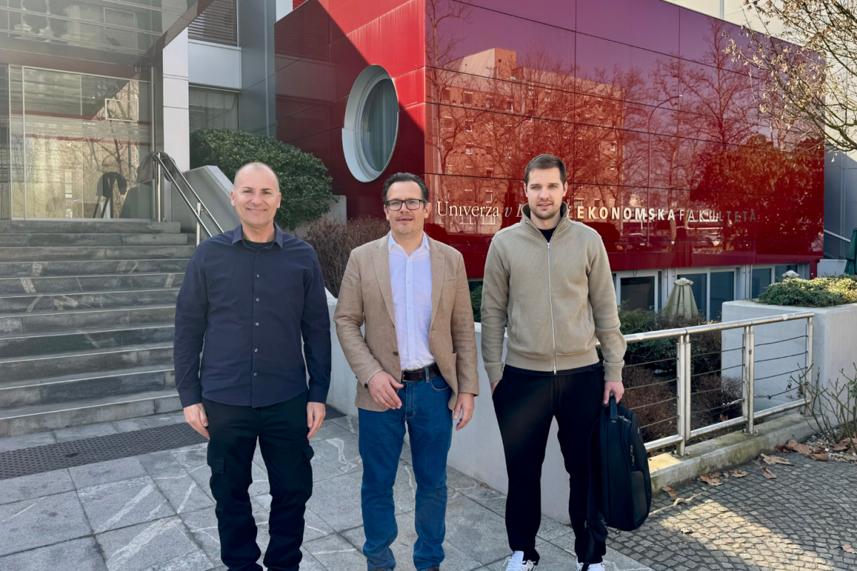 Three men stand in front of a modern building with red accents. The setting is bright and sunny, conveying a professional and cheerful mood.