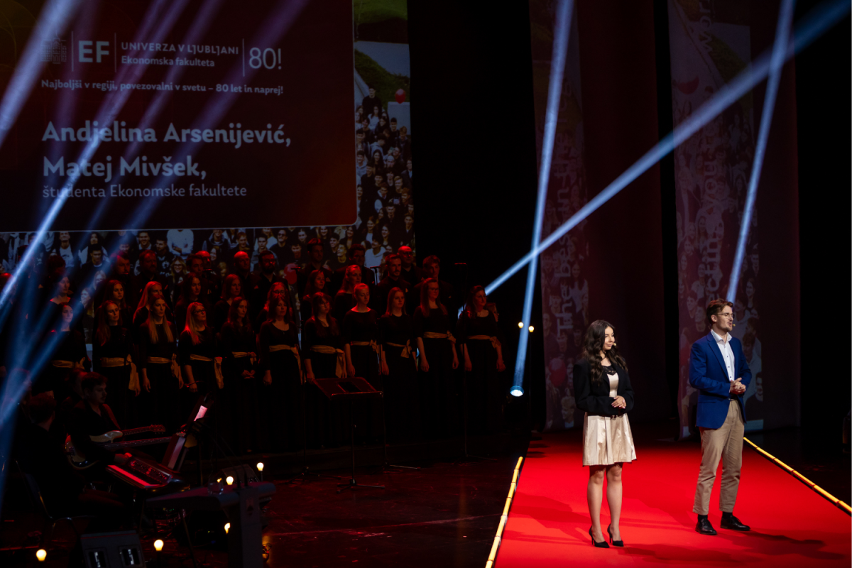 Two presenters stand on a bright red carpet stage under spotlights, with a choir in the background. A large screen displays text in Slovenian. Formal atmosphere.