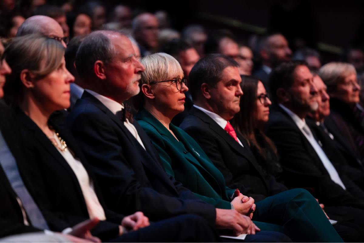 Audience members in formal attire sit attentively in rows at an event. The atmosphere is serious, with soft lighting highlighting their focused expressions.