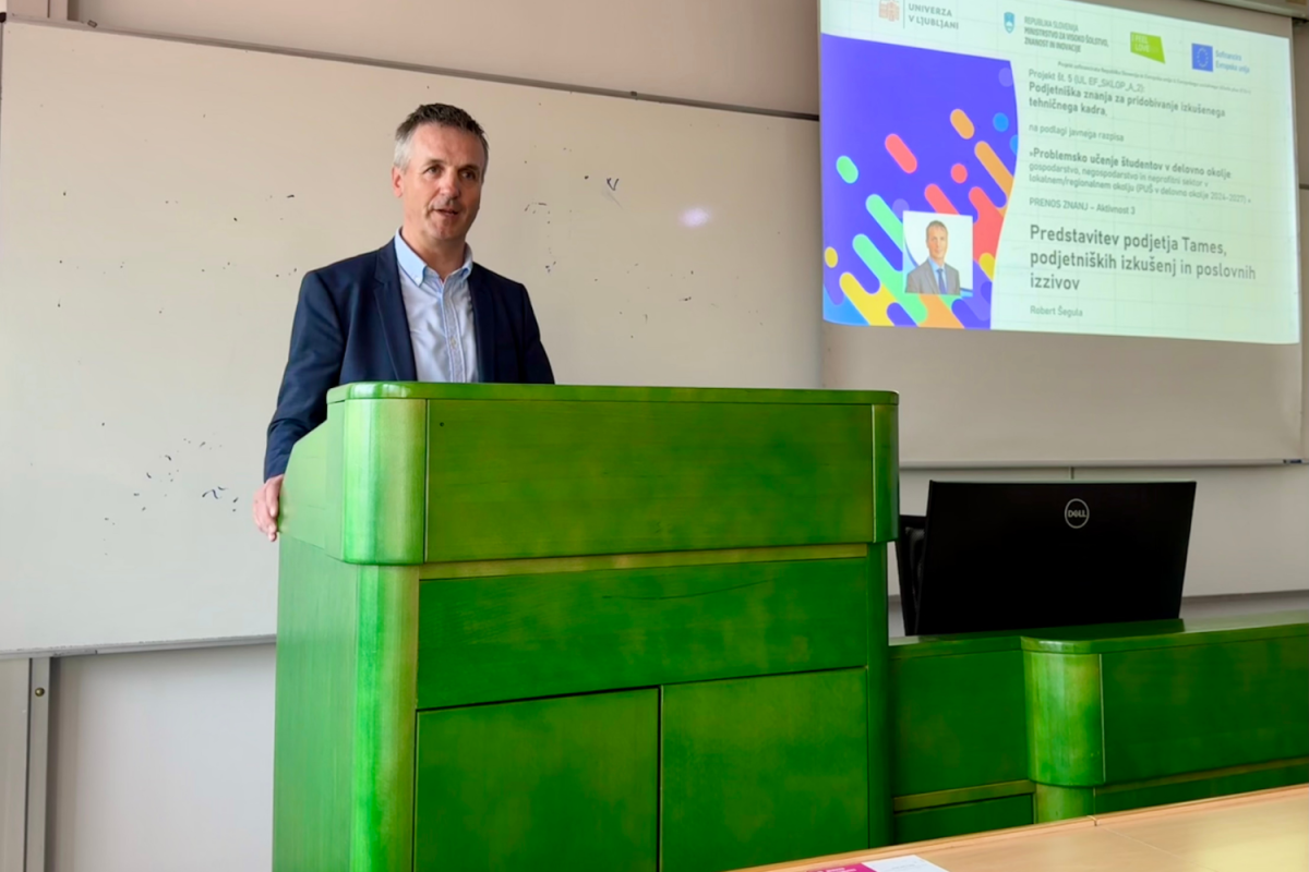 Moški v obleki govori na odru z zeleno leseno oblogo, pred projicirano sliko na tabli v učilnici. Okolje je akademsko in formalno.