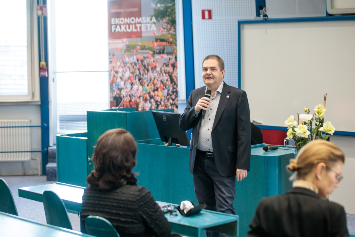 Moški govori v mikrofon v predavalnici, za njim pa je plakat z napisom »Ekonomska Fakulteta«. Udeleženci pozorno poslušajo, kar ustvarja profesionalno in zavzeto vzdušje.