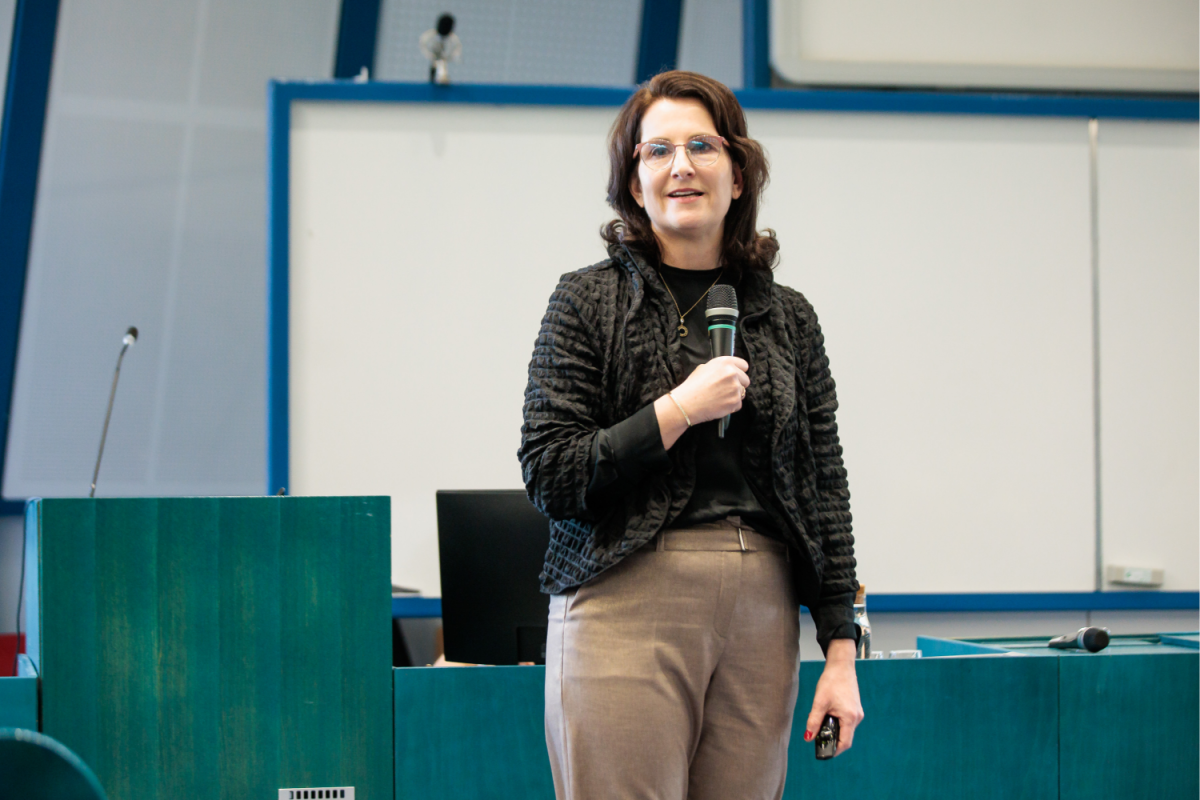 Ženska z očali govori v mikrofon v konferenčni sobi. Nosila je temno majico in bež hlače, izžarevala samozavest in profesionalnost.