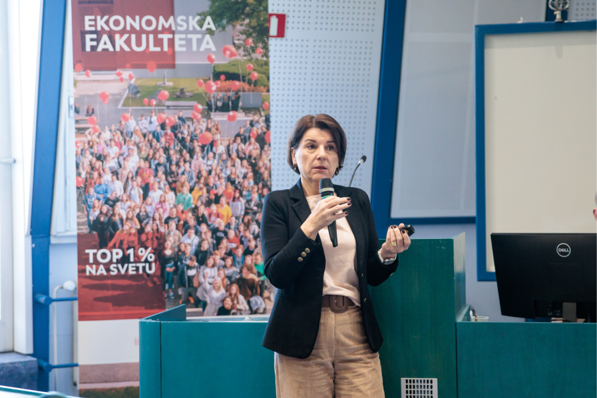 Ženska v poslovni obleki govori v mikrofon med predstavitvijo. Za njo je plakat, na katerem je prikazana množica in napis »Ekonomska Fakulteta, Top 1 % na svetu«.