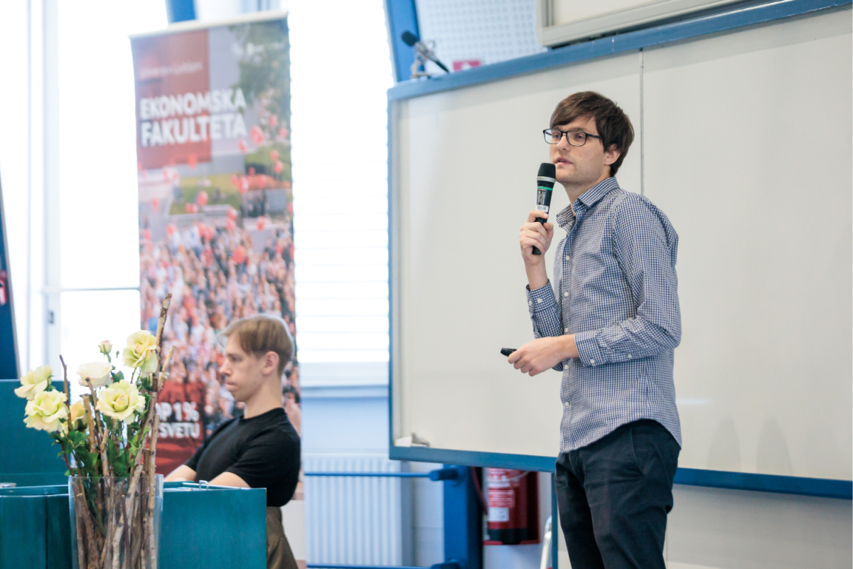 Moški v očalih in karirasti srajci govori v mikrofon v učilnici, stoječ ob tabli. V bližini sedi učenec, v ospredju so cvetje.