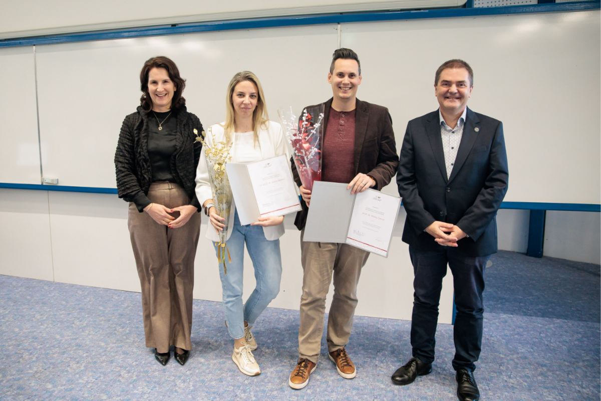 Štirje ljudje stojijo v vrsti v učilnici. Dva v sredini držita certifikate in cvetje, kar nakazuje podelitev nagrad. Razpoloženje je praznično.