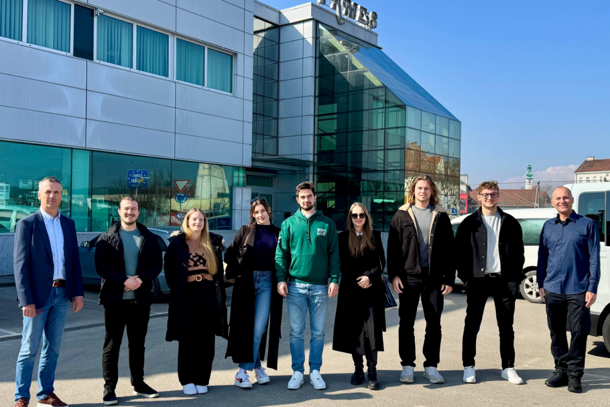 A group of nine people stand smiling in front of a modern building with large glass windows. They're dressed in casual clothing under a clear blue sky.