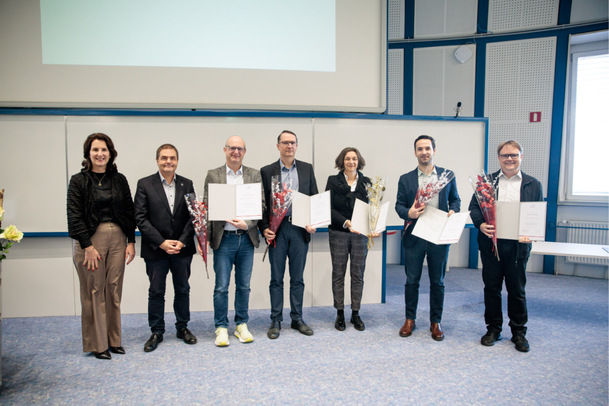 Sedem ljudi stoji nasmejanih v svetli sobi, pet od njih drži certifikate in šopke cvetja, kar nakazuje, da gre za podelitev nagrad. Razpoloženje je praznično.