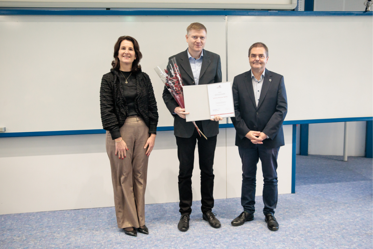 Tri osebe stojijo nasmejane v sobi z belo tablo. Oseba v sredini drži certifikat in cvetje, kar izraža praznično vzdušje.
