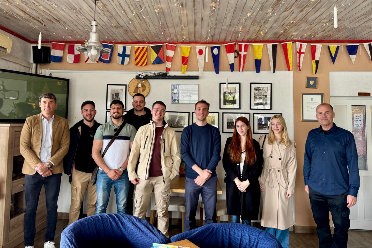 A group of nine people stand in a cozy room with nautical flags overhead. They are smiling, conveying a friendly and relaxed atmosphere.