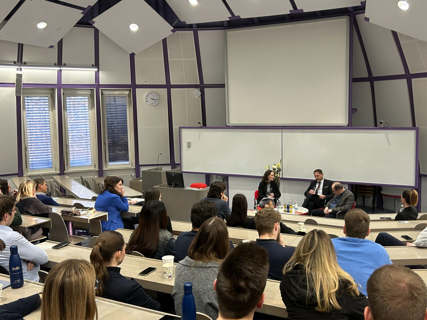 A lecture hall setting featuring a group of people engaged in listening, with a projector screen visible at the front.