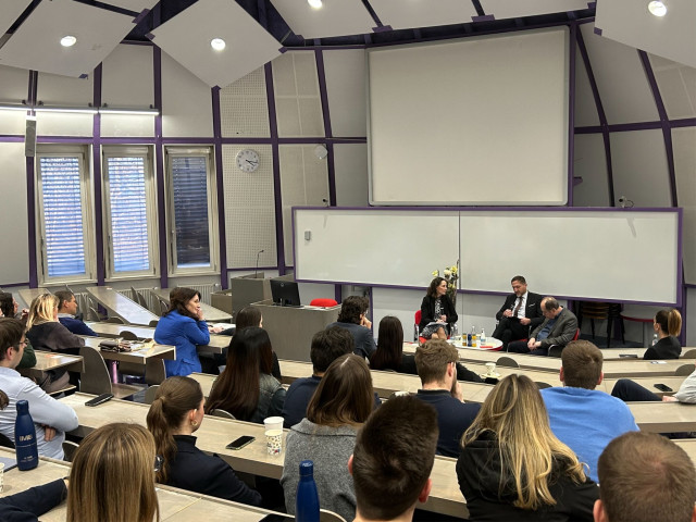 A lecture hall setting featuring a group of people engaged in listening, with a projector screen visible at the front.