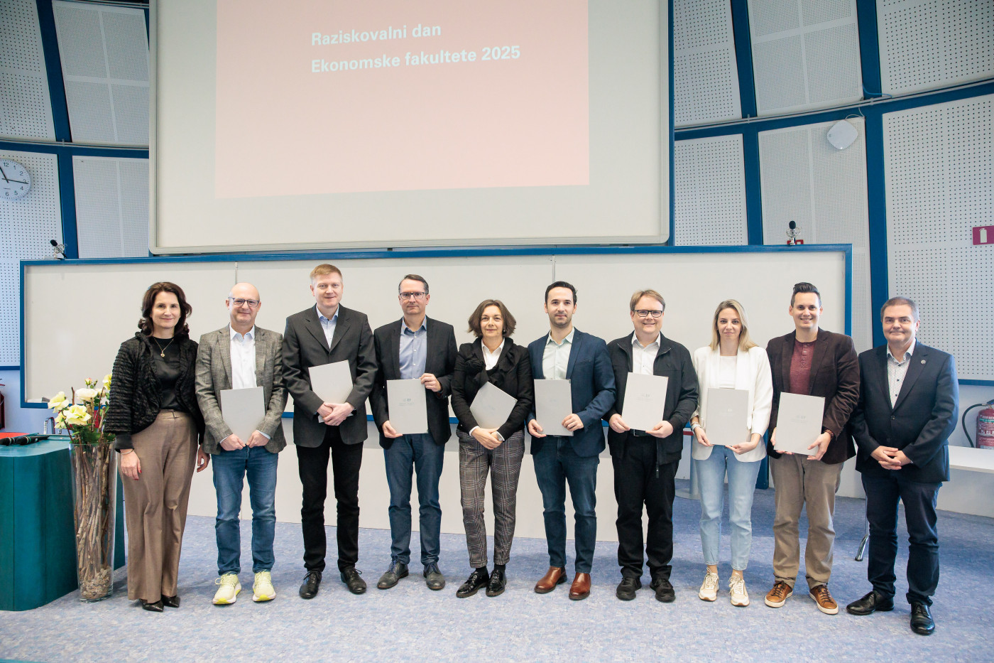 Skupina raziskovalk in raziskovalcev z nagradami pred belo tablo na katedru Plave dvorane Ekonomske fakultete. 