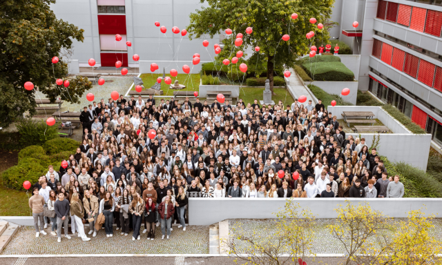 A large group of people standing outdoors, many holding red balloons. The scene is lively with greenery and buildings in the background, creating a joyful atmosphere.