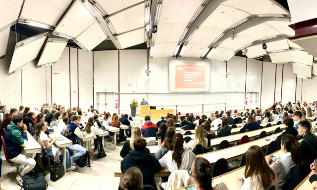 Panoramski pogled na veliko predavalnico, polno pozorno poslušajočih študentov, ki gledajo proti predavatelju na odru. Na zaslonu je prikazana predstavitev.
