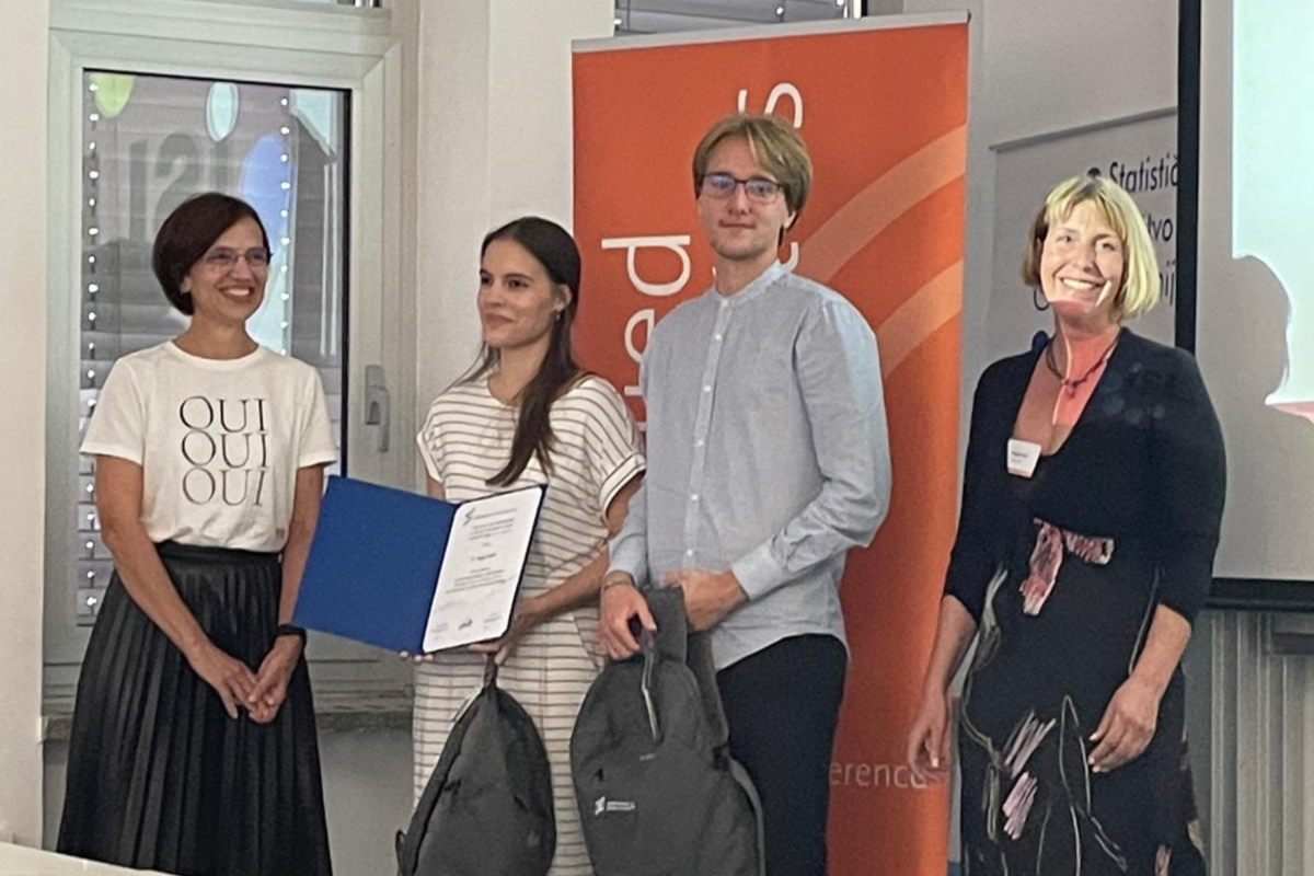 Tri osebe stojijo ob mizi, na kateri je razstavljen certifikat, se smejijo in pogovarjajo.
