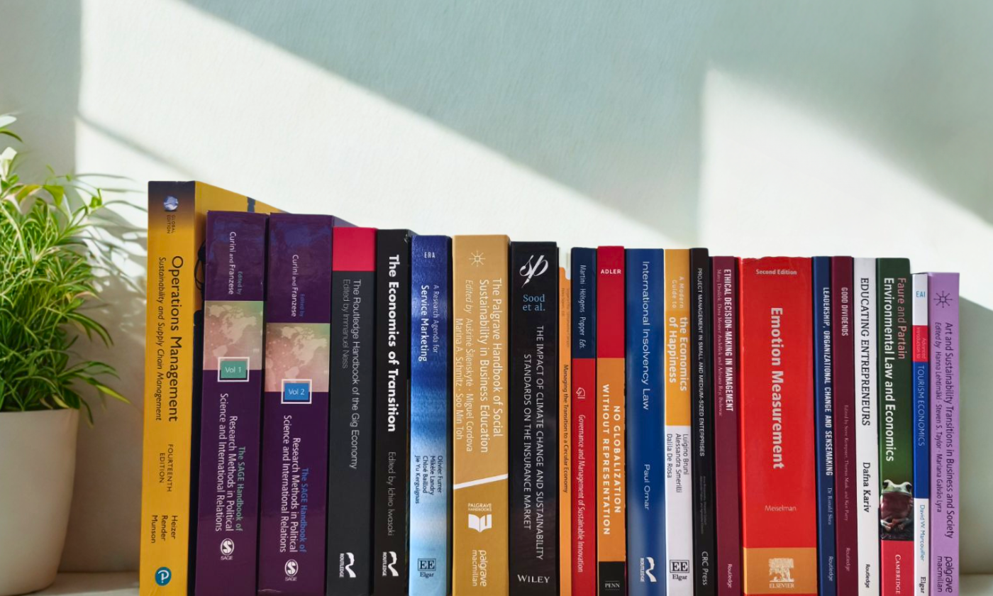 The photograph shows a bookshelf with professional literature in the fields of economics, business, management, and sustainability. On the left side of the image, there is a potted plant.