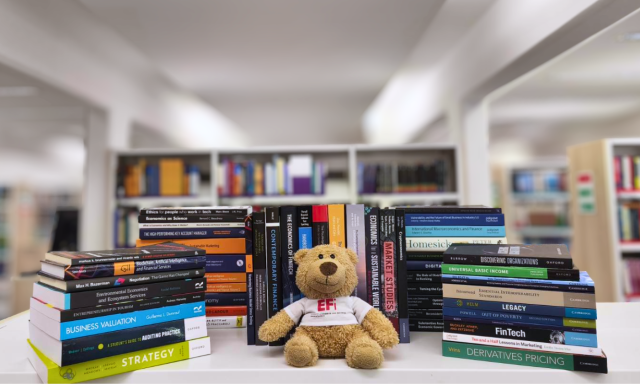 A photograph of new books at the CEK Library displayed on a stand in front of bookshelves; a teddy bear wearing an EFi T-shirt sits among the books.