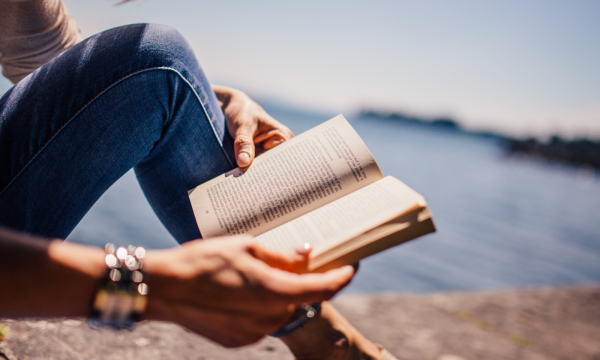 A person sitting by the water reading a book, with summer sunlight illuminating the pages and creating a relaxed atmosphere.