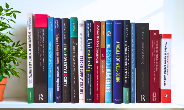 The photograph shows a bookshelf filled with academic books, covering topics such as economics, finance, policy, leadership, and public relations. There is a potted plant on the left side of the image.
