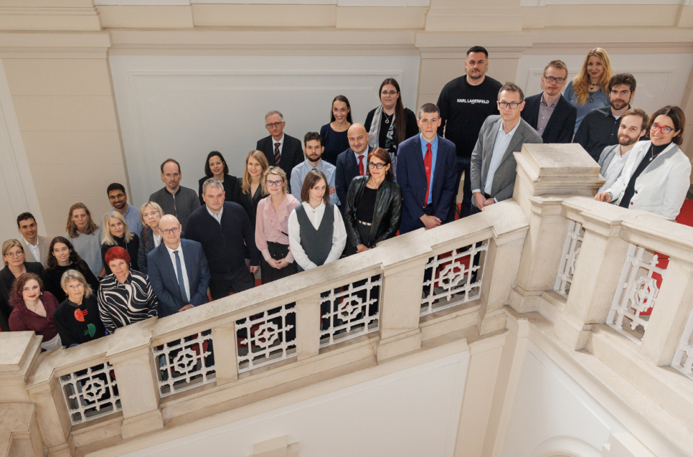 Skupina ljudi stoji na stopnicah znotraj stavbe in gleda v kamero.