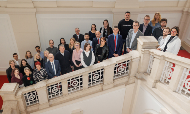 Skupina ljudi stoji na stopnicah znotraj stavbe in gleda v kamero.