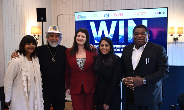 Pet oseb stoji skupaj in se smeji pred zaslonom, na katerem je napisano »WIN« in logotipi sponzorjev dogodka, v dobro osvetljenem notranjem prostoru.