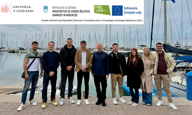 Skupina ljudi stoji nasmejana ob marini, v ozadju pa so vidne jadrnice in jambori. Nebo je oblačno, kar ustvarja sproščeno in neformalno vzdušje.