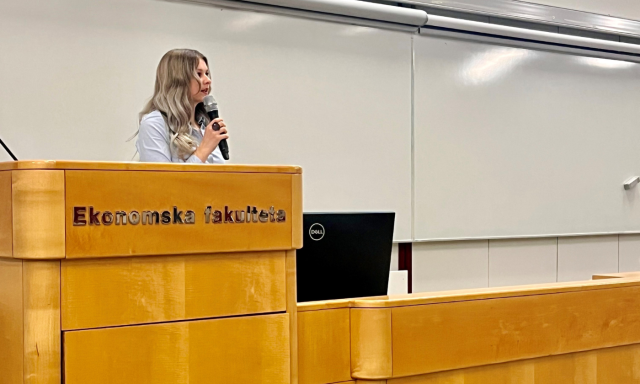 Ženska govori v mikrofon na lesenem odru v predavalnici. Za njo je tabla. Atmosfera je profesionalna in osredotočena.