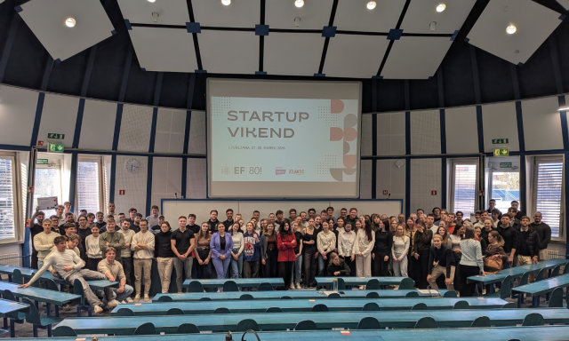 Skupina ljudi se je zbrala v veliki predavalnici in pozirala pred projekcijo na zaslonu.