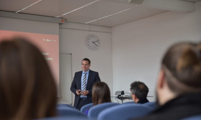 Moški v obleki predava v predavalnici, stoji ob zaslonu in uri, ki kaže 16:13. Udeleženci sedijo na modrih stolih in pozorno poslušajo.