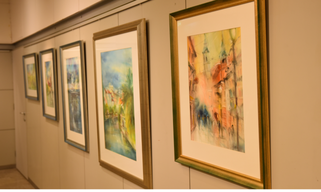 Hodnik umetnostne galerije s petimi okvirjenimi akvareli na bež steni. Umetniška dela prikazujejo abstraktne pokrajine, v katerih se prepletajo živahne modre, zelene in oranžne barve.