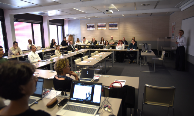 Oseba stoji spredaj v konferenčni dvorani in govori pred pozorno publiko. V dvorani je več prenosnih računalnikov in velik zaslon.