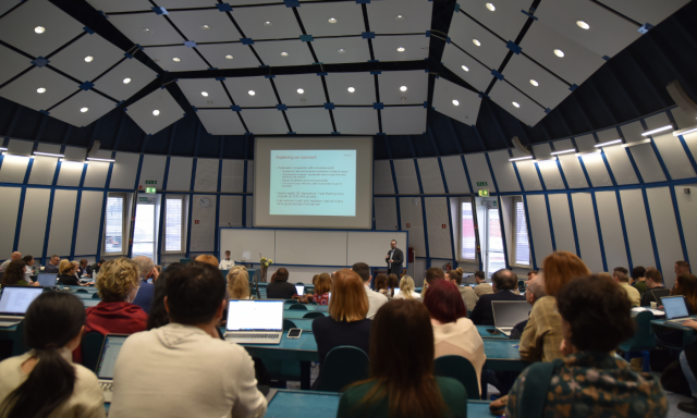 Velika predavalnica z visokim, obloženim stropom. Veliko ljudi sedi z odprtimi prenosnimi računalniki in pozorno spremlja predavatelja, ki stoji spredaj pod projicirano diapozitivno sliko.