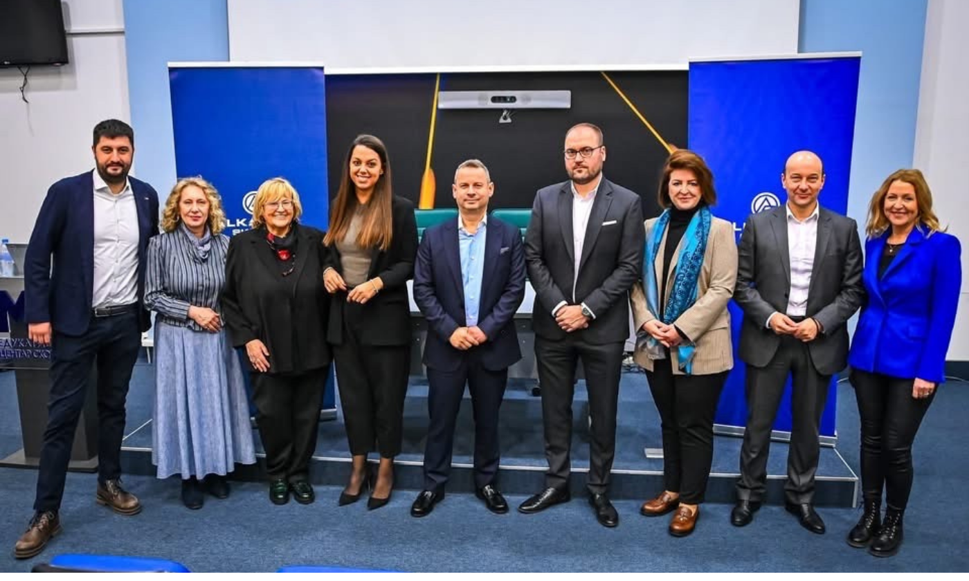 Ten people in business attire stand in a row, posing for a group photo indoors in front of two blue banners and a projection screen.
