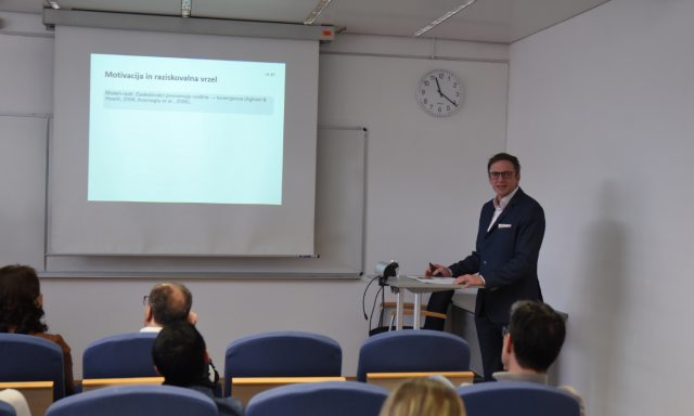 Moški v obleki stoji na odru in predava pred občinstvom, v ozadju pa je vidna projekcija.