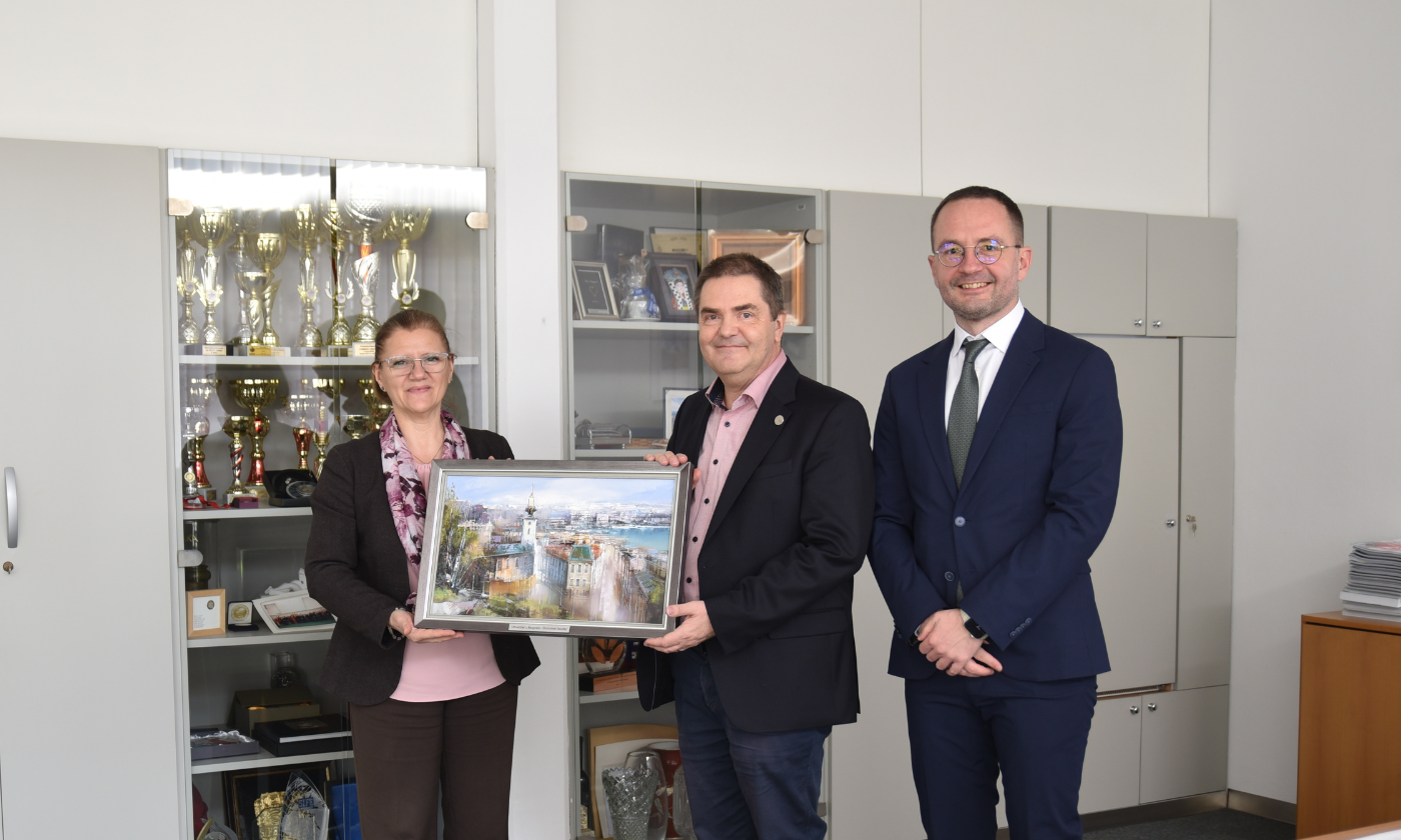 Three people stand in an office; two hold a framed picture, while the third stands beside them. Trophies and awards are visible in cabinets in the background.