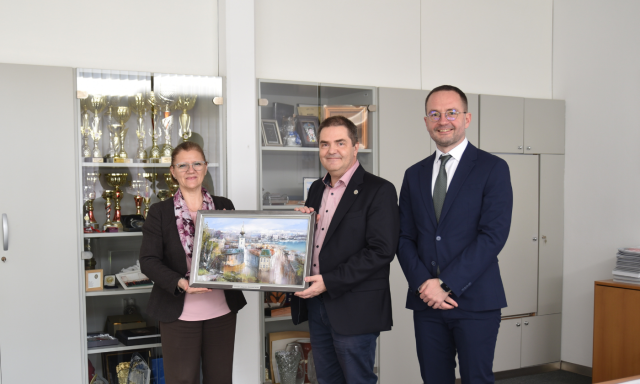 V pisarni stojijo tri osebe; dve držita okvirjeno sliko, tretja pa stoji ob njiju. V omarah v ozadju so vidni pokali in nagrade.