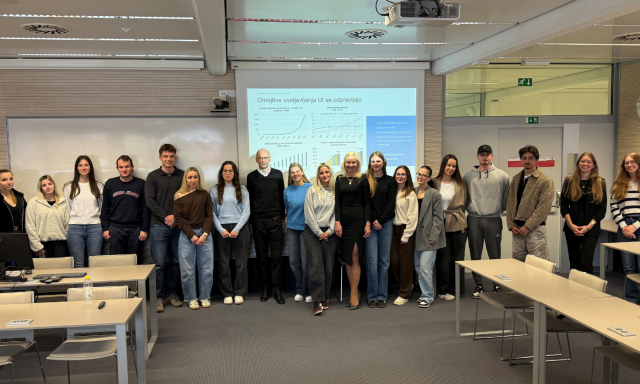 V učilnici pred predstavitvenim diapozitivom stoji raznolika skupina 20 ljudi, ki se nasmiha in ustvarja pozitivno vzdušje sodelovanja.