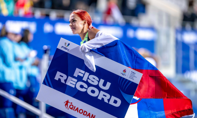 Ženska na prostem z veselim izrazom na obrazu drži plakat z napisom »FISU rekord«.