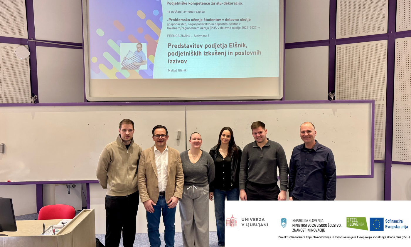 A group of six people stands smiling in front of a classroom screen displaying a presentation title in Slovenian. The setting is a lecture room.