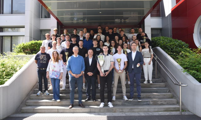 Skupina ljudi s pokali stoji na stopnicah pred stavbo in skupaj proslavlja svoj uspeh.