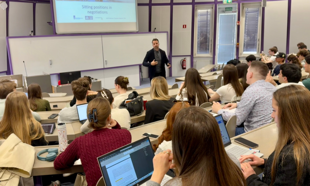 A lecturer in a black suit addresses a classroom of students seated with laptops open, focusing on a presentation about "Sitting positions in negotiations."