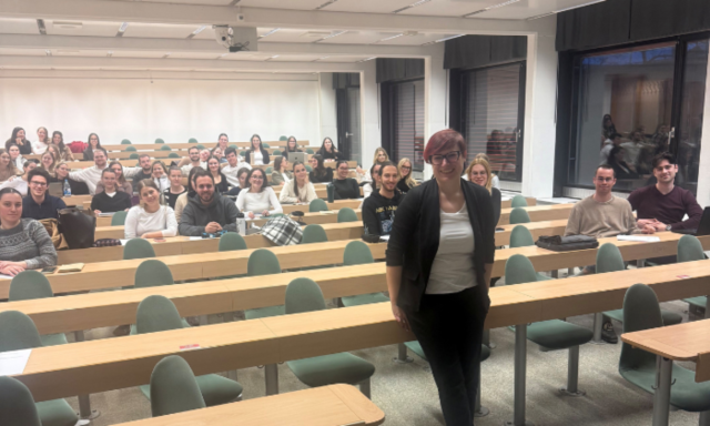 Na čelu univerzitetne predavalnice stoji nasmejana oseba, predavalnica pa je polna pozorno poslušajočih študentov, ki sedijo za vrstami dolgih lesenih miz. Prostor je svetel in prijeten.