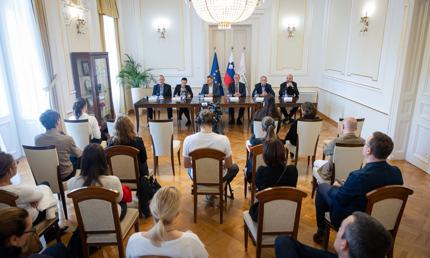 Uradno srečanje v elegantni sobi. Šest oseb sedi za dolgo mizo, obrnjenih proti občinstvu. Za njimi so zastave. Ton je profesionalen in resen.